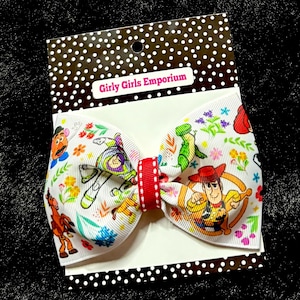 May include: A white hair bow featuring a colorful print of Toy Story characters including Woody, Buzz Lightyear, Mr. Potato Head, and Rex, accented with small floral motifs. The bow has a bright red center band and is attached to a polka dot display card.