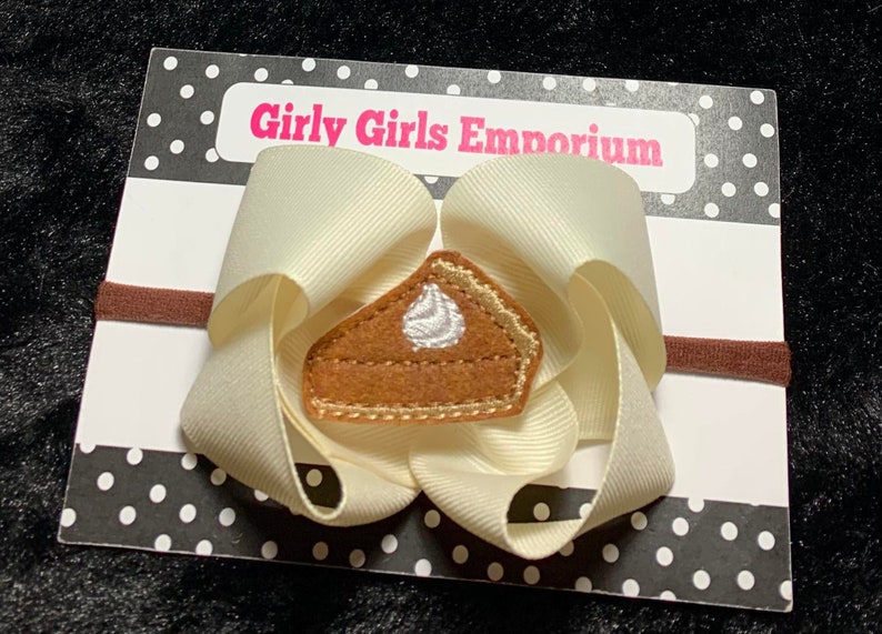 May include: A cream-colored hair bow with a brown and gold felt pie applique. The bow is attached to a brown elastic headband. The card reads "Girly Girls Emporium".
