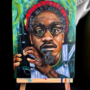 Op de afbeelding: Een acrylverfschilderij op canvas van een man met een donkere huid, met een rode gebreide muts en een ronde zwarte bril. De man heeft een sik en draagt een blauwe overall. Het schilderij wordt tentoongesteld op een klein houten ezel.