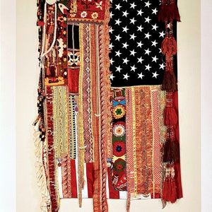 Puede incluir: Una bandera estadounidense roja, blanca y azul hecha de retazos de tela coloridos y borlas. La bandera es un mosaico de diferentes telas, incluyendo rojo, blanco, azul, verde, amarillo y naranja. La bandera está colgada en una pared.