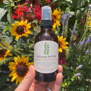 Puede incluir: Una botella de vidrio marrón con una boquilla de pulverización, etiquetada "Pachamama Bug Shield" y "Biching bus bites through nature's vibes". La botella está rodeada de una variedad de flores, incluyendo girasoles amarillos y flores moradas.