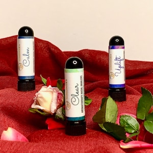 May include: Three aromatherapy nasal inhalers with labels that read "Calm", "Cheer", and "Uplift". The inhalers are on a red fabric background with rose petals and leaves.
