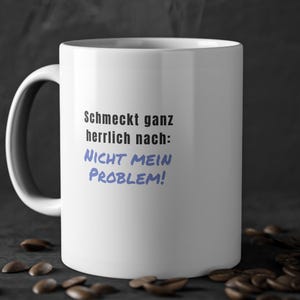 Könnte beinhalten: Eine weiße Keramiktasse mit schwarzem Henkel und dem Text "Schmeckt ganz herrlich nach: NICHT MEIN PROBLEM!" in Schwarz und Blau. Kaffeebohnen sind um die Tasse auf einer dunklen Oberfläche verstreut.