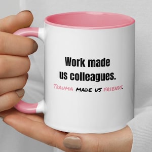 Könnte beinhalten: Weißer Keramikbecher mit rosa Innenraum und Henkel. Der Becher trägt den Text "Work made us colleagues. Trauma made us friends." in schwarzer und rosa Schrift. Der Becher wird von einer Person gehalten.