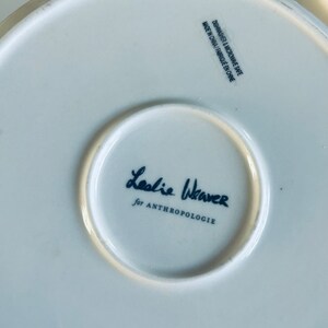 May include: White ceramic plate with a black and white signature that reads "Leslie Weaver for Anthropologie" and the text "Dishwasher & Microwave Safe Made in China/Fabriqu&eacute; en Chine".