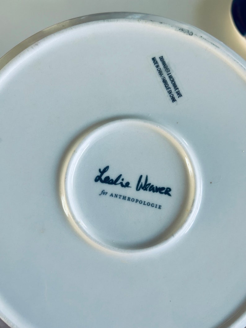 May include: White ceramic plate with a black and white signature that reads "Leslie Weaver for Anthropologie" and the text "Dishwasher & Microwave Safe Made in China/Fabriqu&eacute; en Chine".