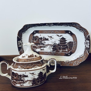 May include: A brown and white porcelain sugar bowl and lid with a Japanese-inspired design. The bowl features a pagoda, bridge, and trees. The matching serving tray has a similar design.