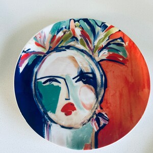 May include: A white ceramic plate with a colorful abstract painting of a woman's face. The painting features a blue background, red and green accents, and a floral design around the woman's head.