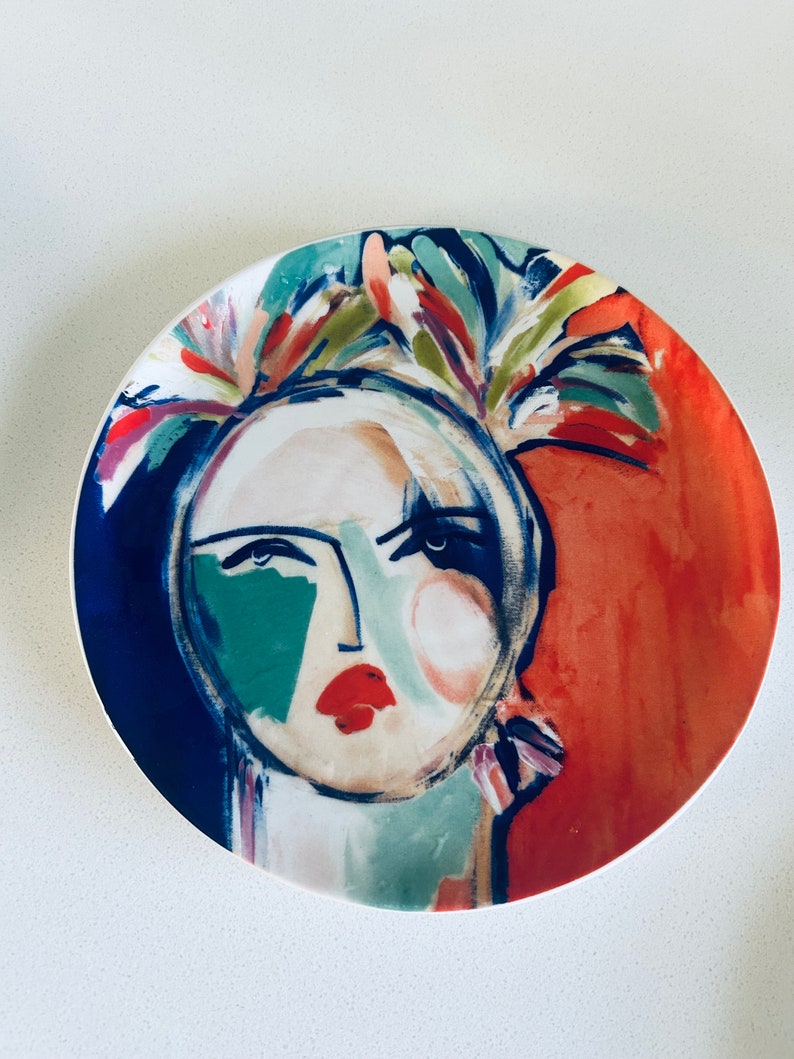 May include: A white ceramic plate with a colorful abstract painting of a woman's face. The painting features a blue background, red and green accents, and a floral design around the woman's head.