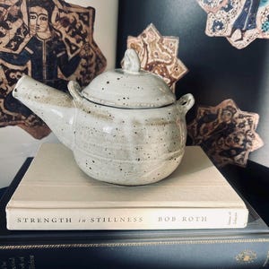 May include: A speckled white ceramic teapot with a lid, sitting on top of two books. The top book has the title "Strength in Stillness" by Bob Roth.