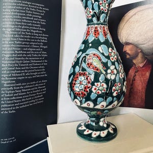 May include: A green ceramic vase with a floral pattern in shades of red, blue, and white. The vase has a round body and a tall, slender neck. It sits on a white surface.
