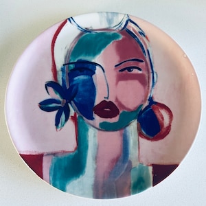 May include: A ceramic plate with a colorful abstract painting of a woman's face. The painting features shades of pink, blue, green, and white.