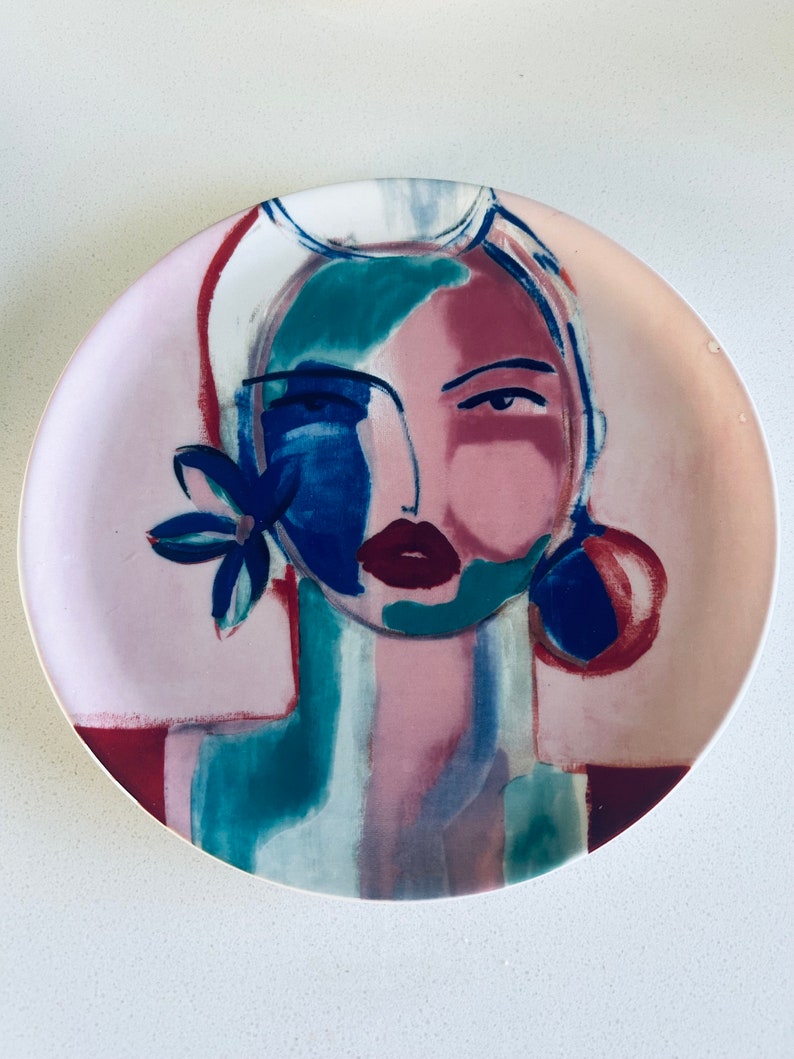 May include: A ceramic plate with a colorful abstract painting of a woman's face. The painting features shades of pink, blue, green, and white.