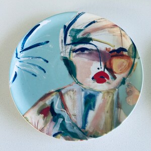 May include: A blue ceramic plate with a colorful abstract painting of a person's face. The painting features a woman with red lips, blue eyes, and a yellow background.