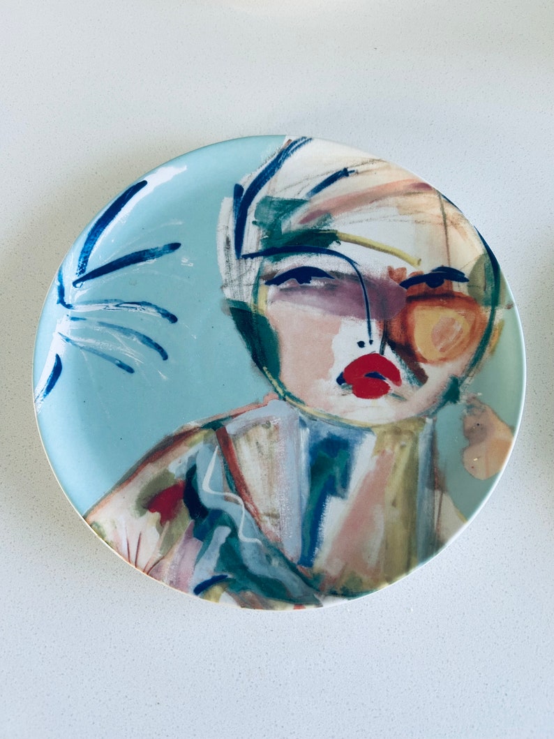 May include: A blue ceramic plate with a colorful abstract painting of a person's face. The painting features a woman with red lips, blue eyes, and a yellow background.