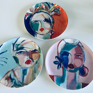 May include: Three decorative ceramic plates with abstract painted designs of faces. Each plate features a different color scheme and facial expression. The plates are round and have a white background.