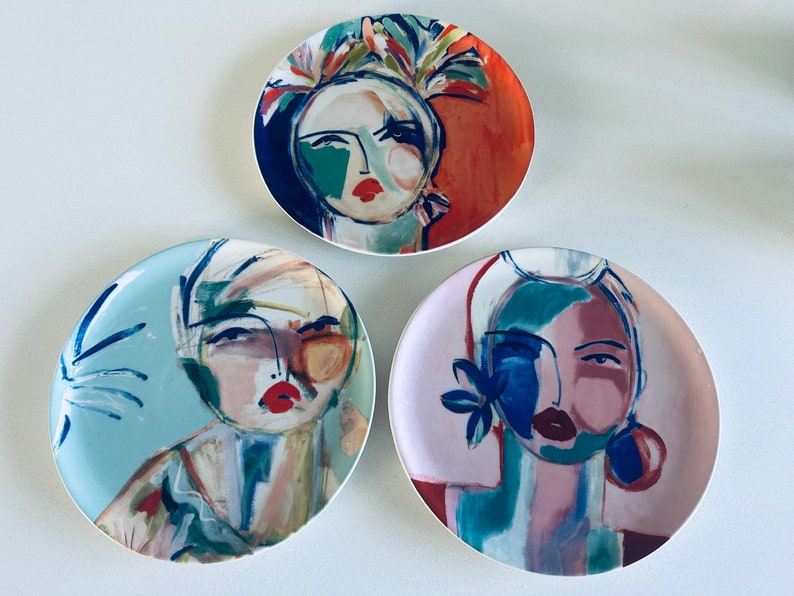 May include: Three decorative ceramic plates with abstract painted designs of faces. Each plate features a different color scheme and facial expression. The plates are round and have a white background.