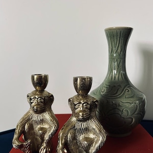 May include: Two gold monkey candle holders with a green glazed ceramic vase in the background. The vase has a floral design.
