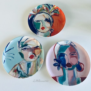 May include: Three round ceramic plates with colorful abstract paintings of women's faces. Each plate features a different color scheme and design.