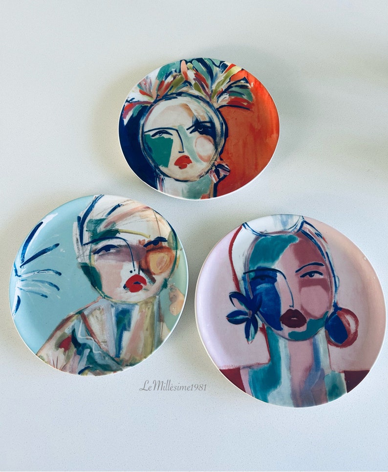 May include: Three round ceramic plates with colorful abstract paintings of women's faces. Each plate features a different color scheme and design.