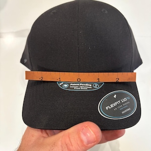 May include: A black Flexfit 110 hat with a curved brim. The hat has a structured front and an adjustable closure. The text "Patent Pending" and "Thinner | Stitchless Front Seam" is visible. The hat measures approximately 8 inches across.