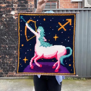 May include: Crocheted wall hanging featuring the Sagittarius zodiac sign. The design includes a centaur with a bow and arrow, set against a dark blue background with stars and the Sagittarius symbol. The border is yellow.