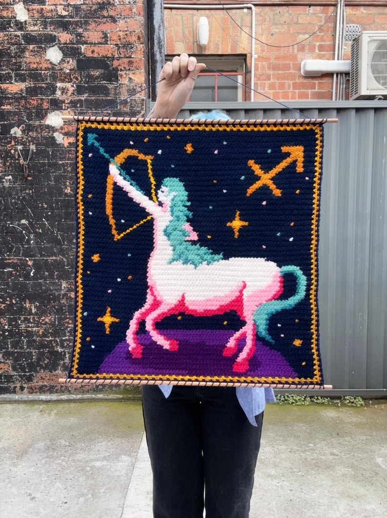 May include: Crocheted wall hanging featuring the Sagittarius zodiac sign. The design includes a centaur with a bow and arrow, set against a dark blue background with stars and the Sagittarius symbol. The border is yellow.
