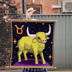 May include: A handmade wall hanging featuring a crocheted image of the Taurus zodiac sign. The design includes a yellow bull on a purple platform against a dark blue background with stars. The bull has pink horns and a pink nose. The piece is framed with wooden dowels.