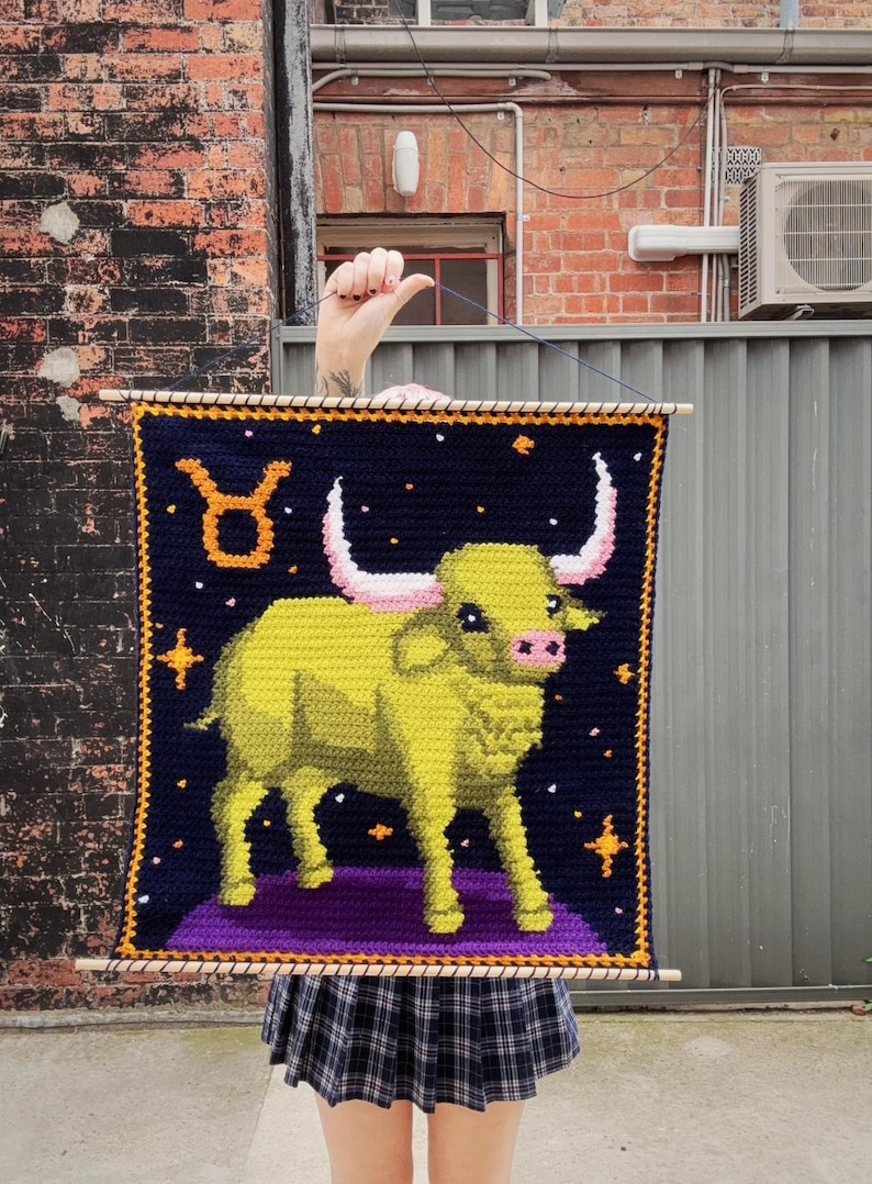 May include: A handmade wall hanging featuring a crocheted image of the Taurus zodiac sign. The design includes a yellow bull on a purple platform against a dark blue background with stars. The bull has pink horns and a pink nose. The piece is framed with wooden dowels.