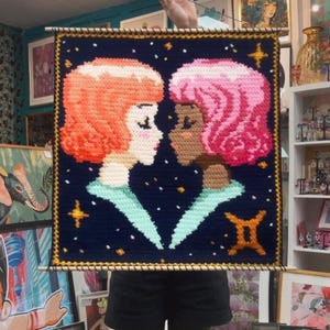 May include: A handmade wall hanging featuring two stylized female figures facing each other. One figure has orange hair, the other pink, against a dark blue background with stars and a Gemini symbol. The artwork is framed with a wooden dowel.
