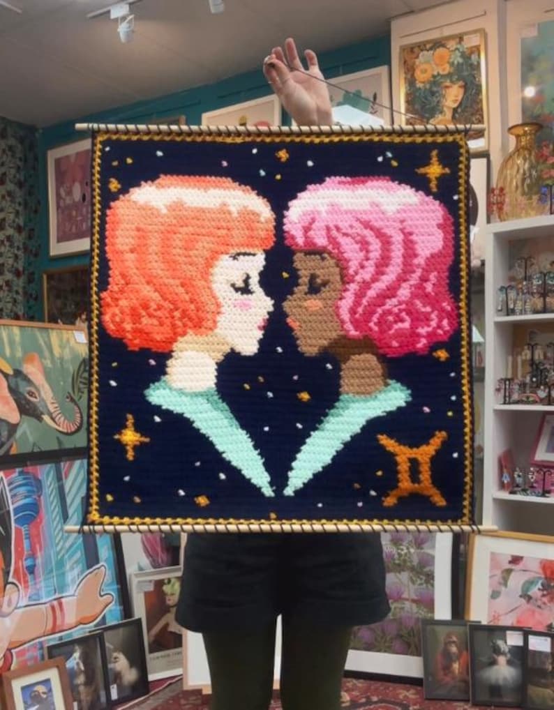 May include: A handmade wall hanging featuring two stylized female figures facing each other. One figure has orange hair, the other pink, against a dark blue background with stars and a Gemini symbol. The artwork is framed with a wooden dowel.