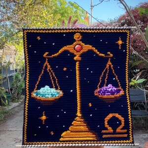 May include: A navy blue crochet wall hanging featuring the Libra zodiac symbol. The design includes a golden scale with a red gem, holding turquoise and purple piles. The background has golden stars and a golden border. The piece is held by a wooden dowel.