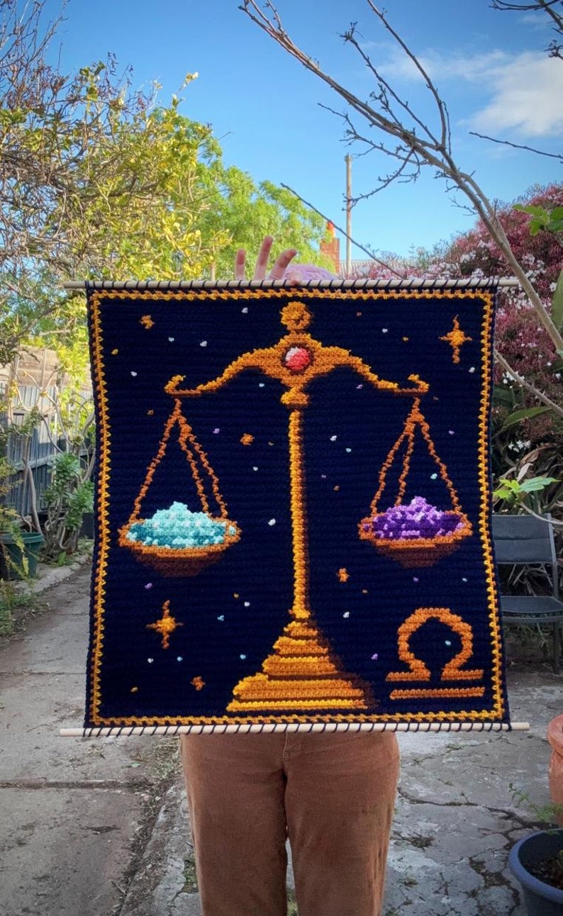 May include: A navy blue crochet wall hanging featuring the Libra zodiac symbol. The design includes a golden scale with a red gem, holding turquoise and purple piles. The background has golden stars and a golden border. The piece is held by a wooden dowel.