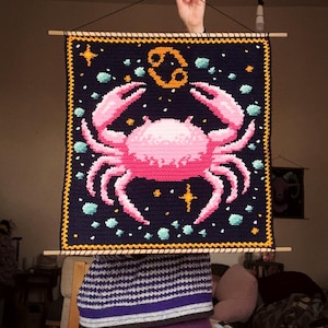 May include: A handmade, square wall hanging featuring a pink crab design on a dark blue background. The crab is surrounded by stars and turquoise accents. The zodiac symbol for Cancer is in the upper center. The piece is framed with a yellow border and wooden dowels.