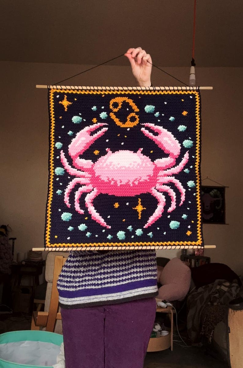 May include: A handmade, square wall hanging featuring a pink crab design on a dark blue background. The crab is surrounded by stars and turquoise accents. The zodiac symbol for Cancer is in the upper center. The piece is framed with a yellow border and wooden dowels.