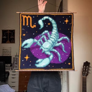 May include: Crocheted wall hanging featuring a scorpion zodiac design. The artwork showcases a teal scorpion on a purple background, set against a navy blue field with gold stars and trim. The word "♏" is in gold.
