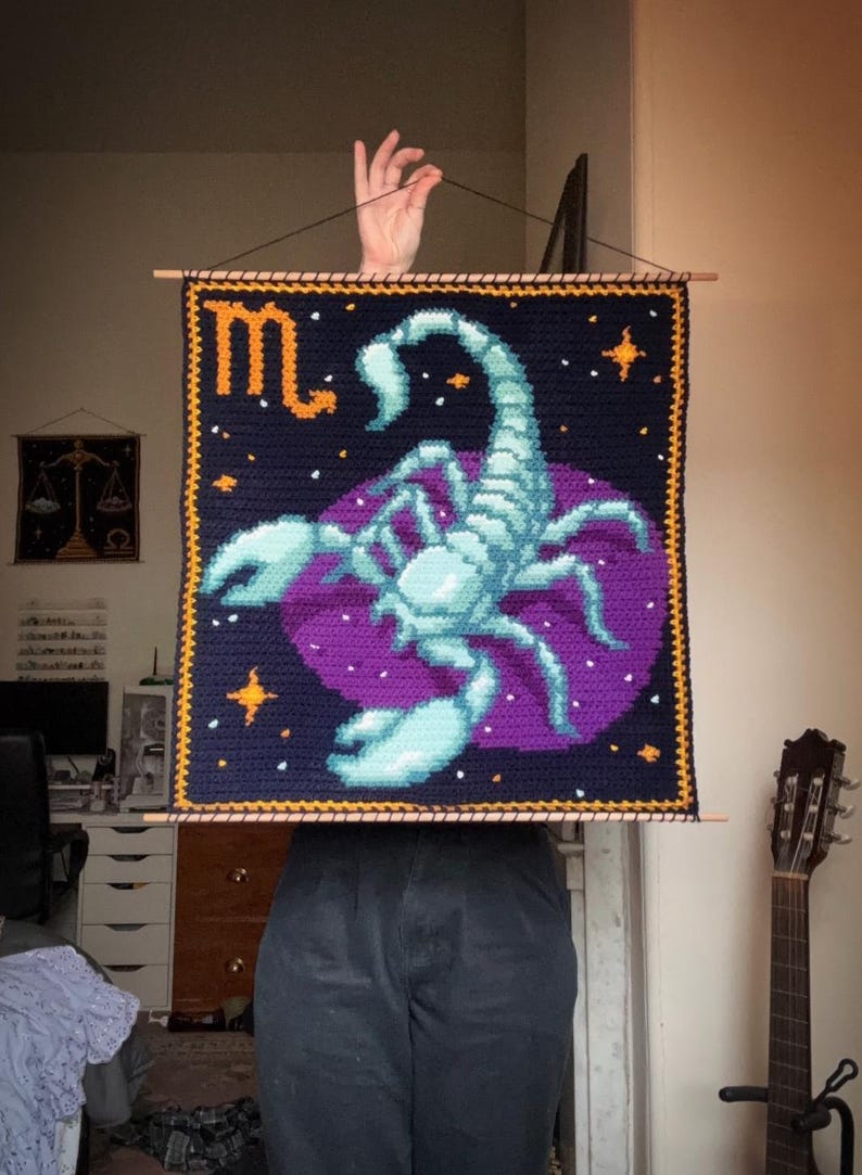 May include: Crocheted wall hanging featuring a scorpion zodiac design. The artwork showcases a teal scorpion on a purple background, set against a navy blue field with gold stars and trim. The word "♏" is in gold.