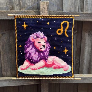 May include: A handmade tapestry featuring a lion, the Leo zodiac sign. The lion is depicted in pink and purple, set against a dark blue background with stars and a gold border. The tapestry is hung with wooden dowels.