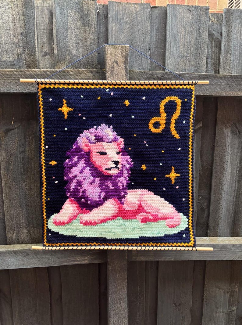 May include: A handmade tapestry featuring a lion, the Leo zodiac sign. The lion is depicted in pink and purple, set against a dark blue background with stars and a gold border. The tapestry is hung with wooden dowels.