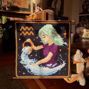 May include: A handmade crochet wall hanging featuring the Aquarius zodiac sign. The artwork depicts a woman with flowing green hair, pouring water from a golden vessel. The background is dark blue with stars, and the border is orange.