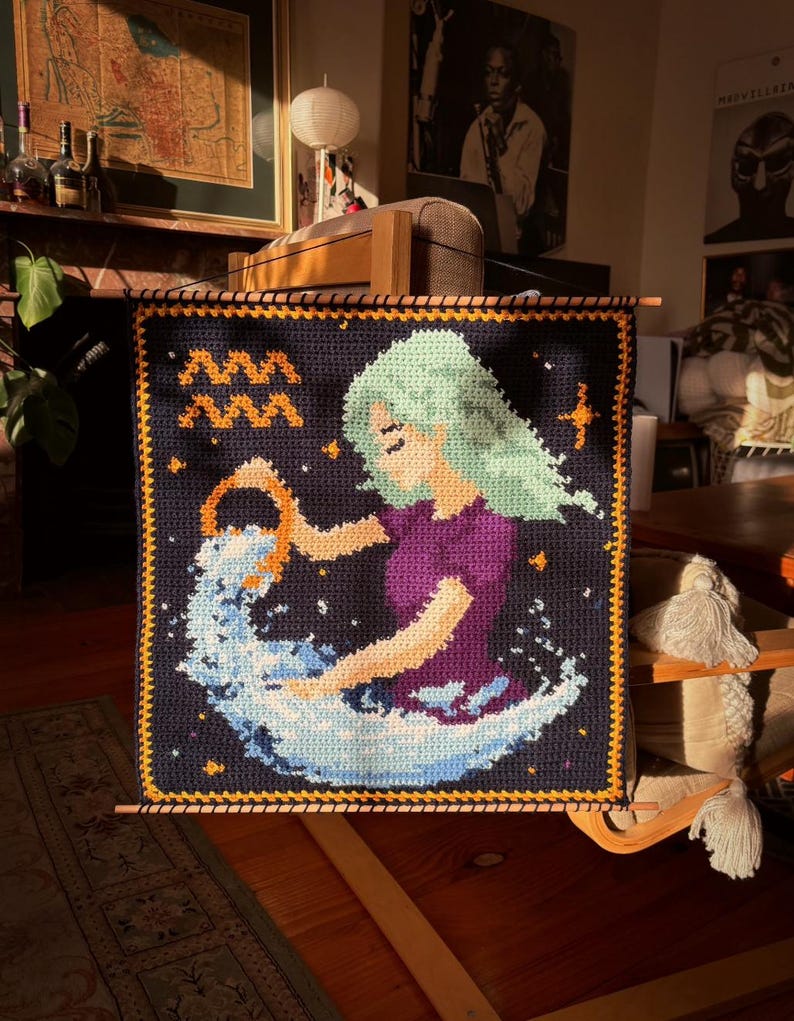 May include: A handmade crochet wall hanging featuring the Aquarius zodiac sign. The artwork depicts a woman with flowing green hair, pouring water from a golden vessel. The background is dark blue with stars, and the border is orange.