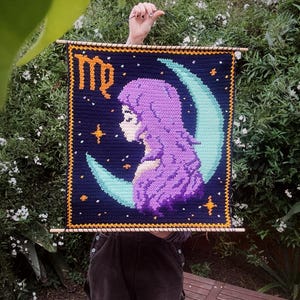 May include: A handmade crochet wall hanging featuring a Virgo zodiac design. The artwork depicts a woman's profile in purple, set against a dark blue background with a crescent moon and stars. The word "Virgo" is written in orange.