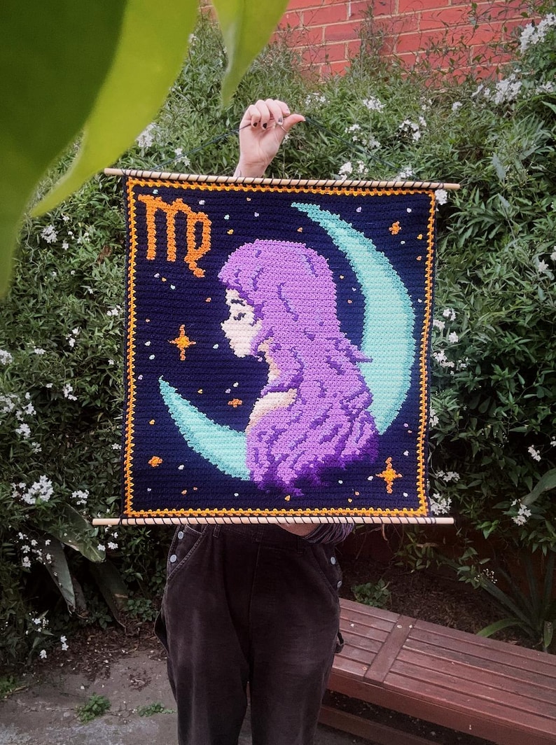 May include: A handmade crochet wall hanging featuring a Virgo zodiac design. The artwork depicts a woman's profile in purple, set against a dark blue background with a crescent moon and stars. The word "Virgo" is written in orange.