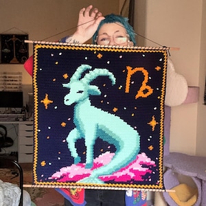May include: A handmade, square tapestry featuring a light blue goat, the Capricorn zodiac symbol, and stars on a dark blue background. The tapestry is framed with a yellow border and wooden dowels.