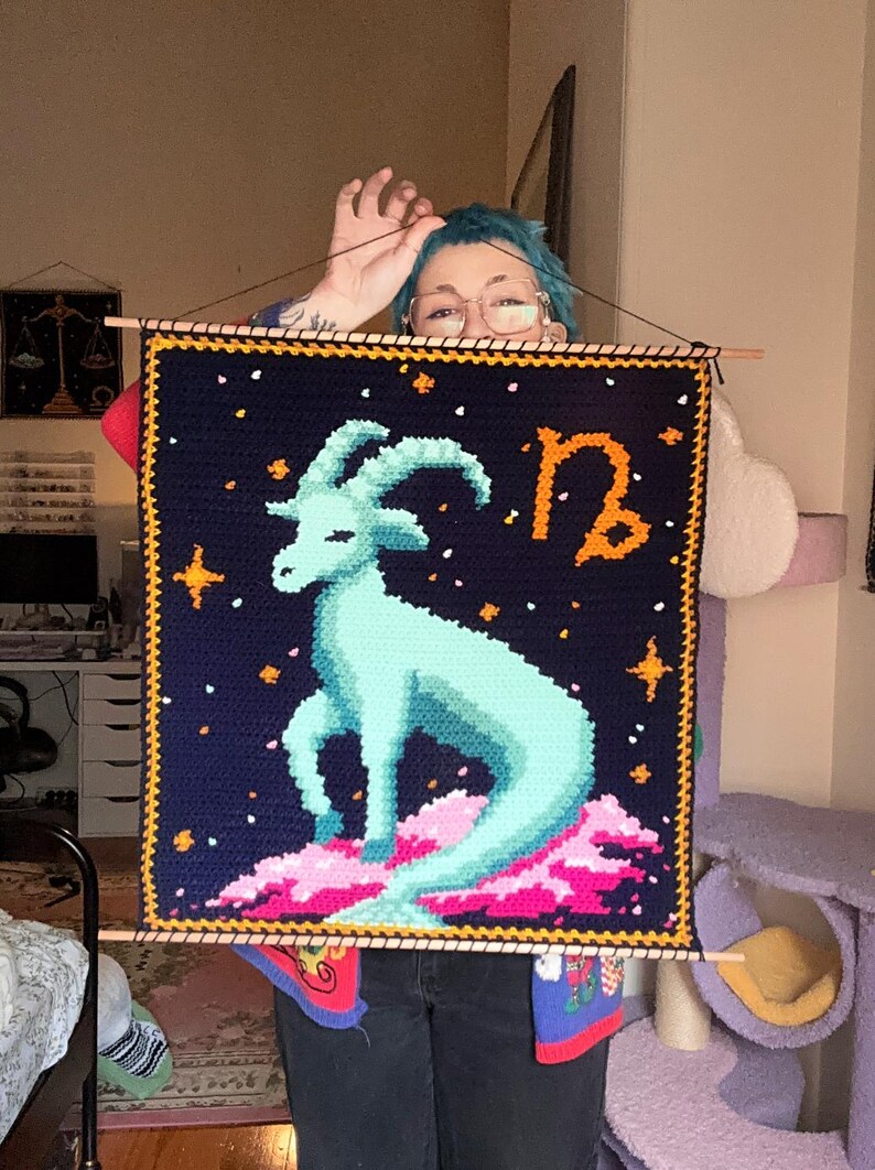 May include: A handmade, square tapestry featuring a light blue goat, the Capricorn zodiac symbol, and stars on a dark blue background. The tapestry is framed with a yellow border and wooden dowels.