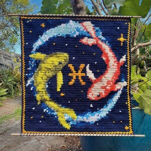May include: Crocheted wall hanging featuring a Pisces design. The artwork depicts two fish in yellow and red, swimming in a blue and white swirl against a dark blue background. The Pisces symbol is in the center. The piece is framed with a yellow border.