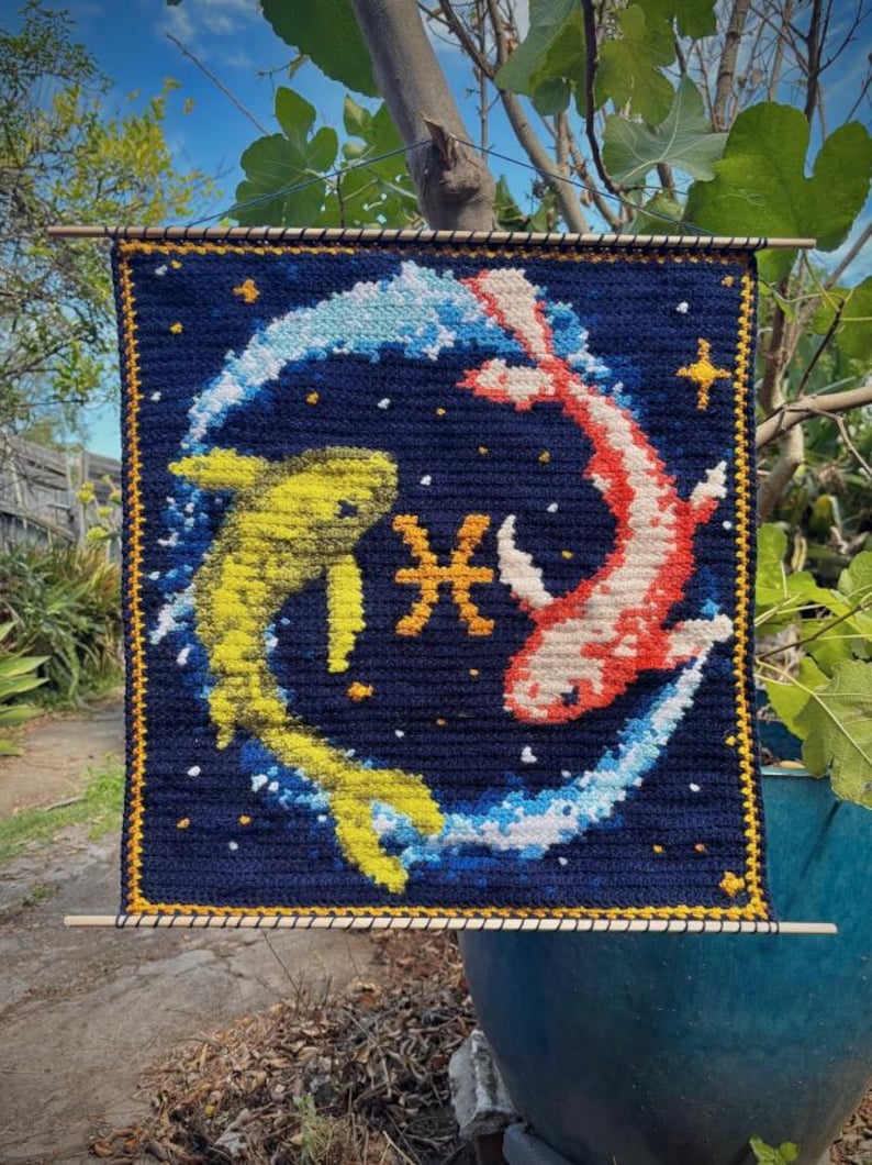 May include: Crocheted wall hanging featuring a Pisces design. The artwork depicts two fish in yellow and red, swimming in a blue and white swirl against a dark blue background. The Pisces symbol is in the center. The piece is framed with a yellow border.