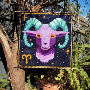 May include: Crocheted wall hanging featuring the Aries zodiac sign. The design includes a purple ram's head with teal horns, set against a navy blue background with stars and a gold border. The Aries symbol is also included.