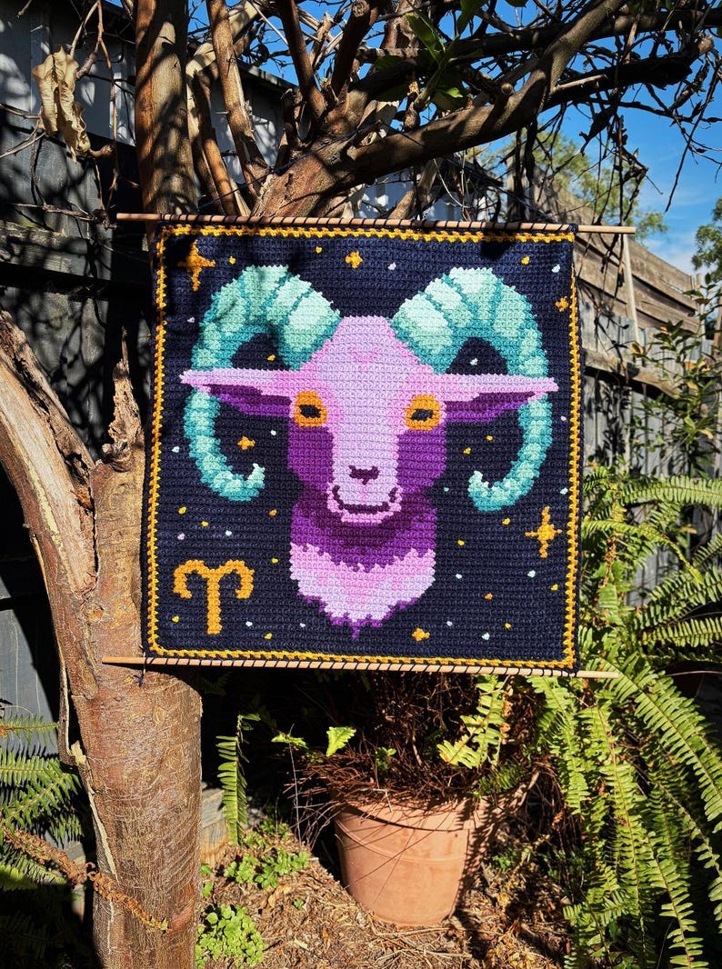 May include: Crocheted wall hanging featuring the Aries zodiac sign. The design includes a purple ram's head with teal horns, set against a navy blue background with stars and a gold border. The Aries symbol is also included.