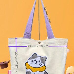 May include: Cream-colored tote bag with a cat design. The bag has purple handles and a button closure. The cat is white and gray with a yellow collar. The bag is 10.63 inches wide, 7.87 inches tall, and 3.54 inches deep.